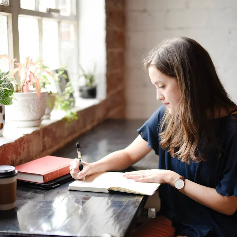 An adult writing in a notebook by a sunlit window.